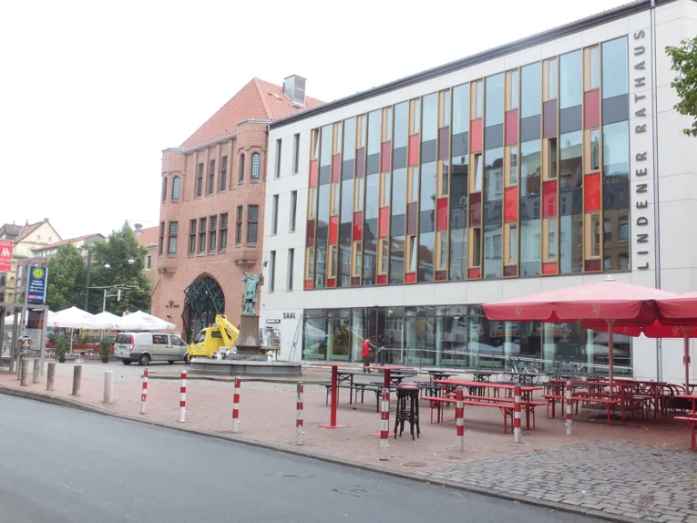 Eine Straße, dahinter ein Platz mit einem Brunnen, einem Sonnenschirm und Bierzeltgarnituren. Dahinter ein Gebäude mit gläserner Fassade und danaben ein Backsteingebäude.
